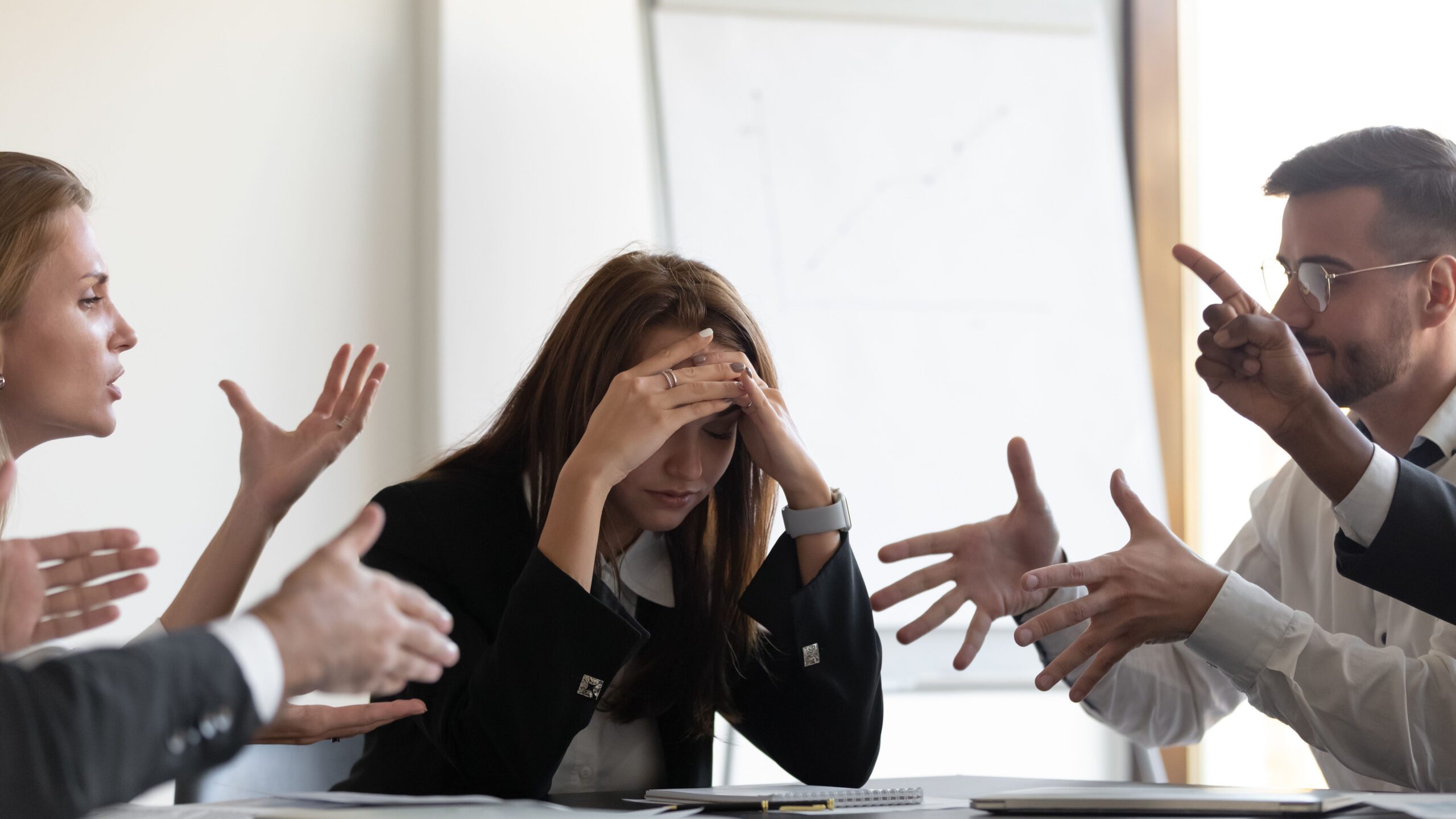 A frustrated woman has her head in her hands with quarreling colleagues at a business meeting