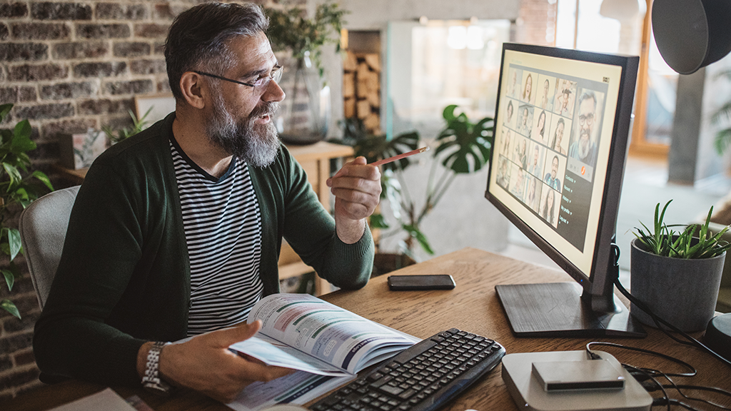 A man with gray beard sits at a desk with a textbook in his hands as he speaks at the computer on a virtual meeting