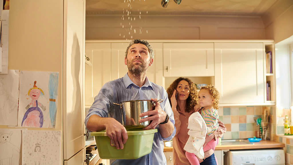 A man holds a tub as water falls from the ceiling with a woman on the phone in the background