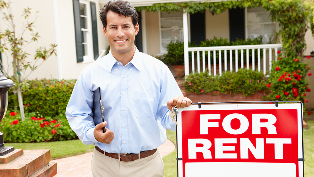 A man stands next to a for rent sign and holds keys and a clipboard