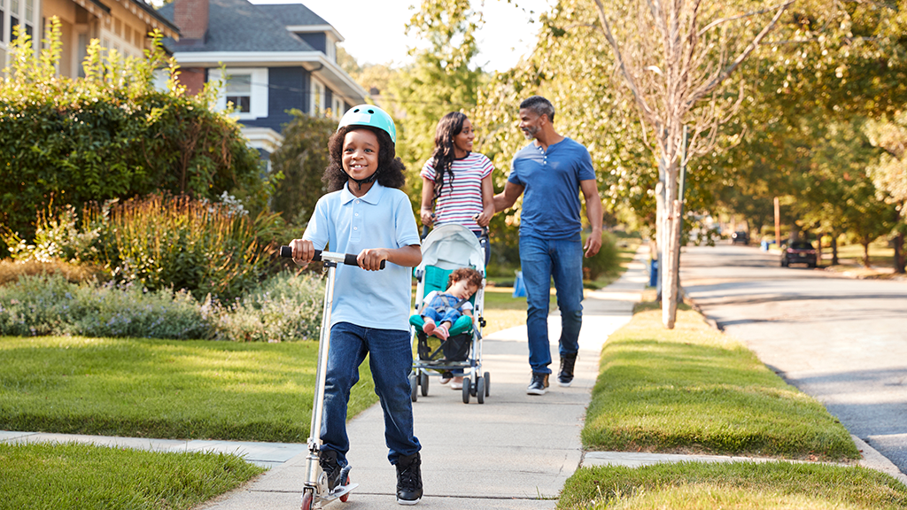 A young child rides a scooter through a suburban sidewalk as his parents push another child in a stroller