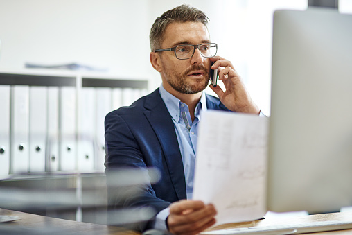 Mature businessman using phone and computer while reading a document at a desk